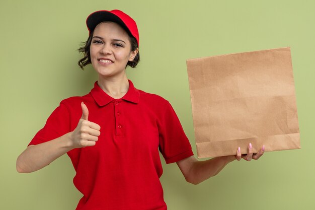 Smiling young pretty delivery woman holding paper food packaging and thumbing up