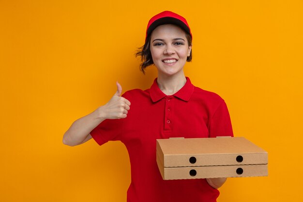 Smiling young pretty delivery woman holding pizza boxes and thumbing up 