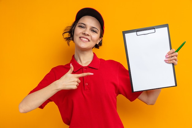 Smiling young pretty delivery woman holding and pointing at clipboard 