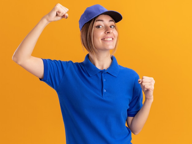 Бесплатное фото Улыбающаяся молодая красивая женщина-доставщик в униформе стоит с поднятыми кулаками, изолированными на оранжевой стене