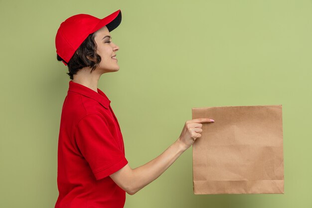 Smiling young pretty delivery woman standing sideways holding paper food packaging 