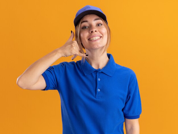 Smiling young pretty delivery woman in uniform gestures call me hand sign isolated on orange wall