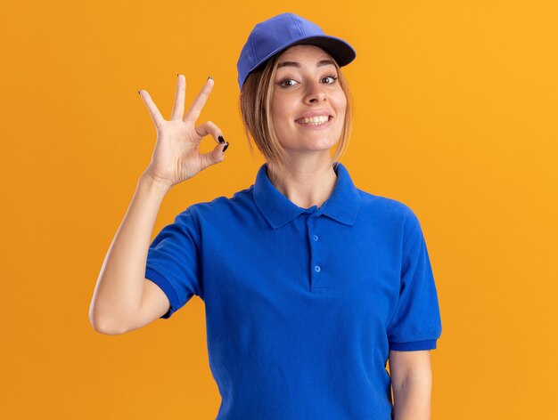Smiling young pretty delivery woman in uniform gestures ok hand sign isolated on orange wall