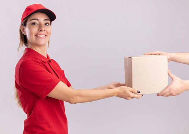 Улыбающаяся молодая красивая женщина-доставщик в униформе дает картонную коробку кому-то, смотрящему вперед, изолированному на белой стене
