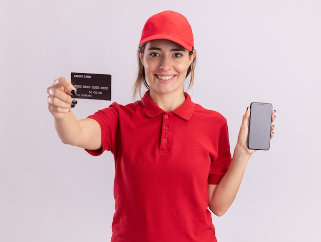 Smiling young pretty delivery woman in uniform holding credit card and phone isolated on white wall