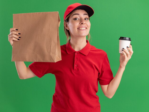 smiling young pretty delivery woman in uniform holds paper cup and paper package isolated on green wall