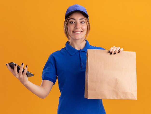 Smiling young pretty delivery woman in uniform holds paper package and phone isolated on orange wall