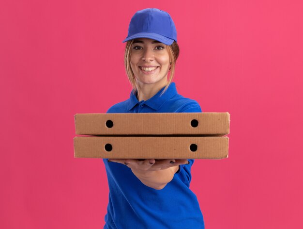 제복을 입은 웃는 젊은 예쁜 배달 여자는 피자 상자를 보유하고 분홍색 벽에 고립 된 정면에 보인다