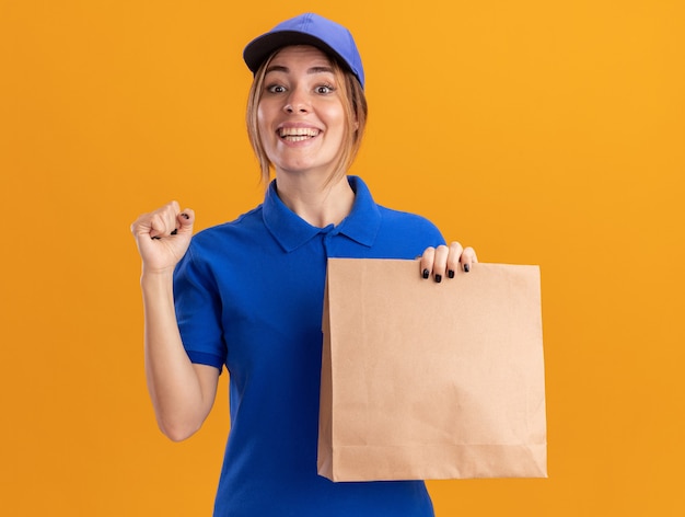 Улыбающаяся молодая симпатичная женщина-доставщик в униформе держит кулак и держит бумажный пакет, изолированный на оранжевой стене