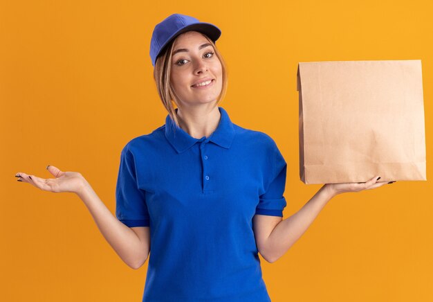 Улыбающаяся молодая красивая женщина-доставщик в униформе держит руку открытой и держит бумажный пакет изолированным