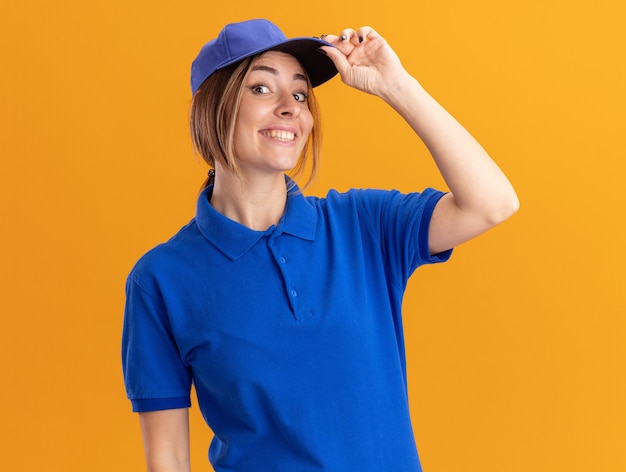 Smiling young pretty delivery woman in uniform puts hand on cap isolated on orange wall