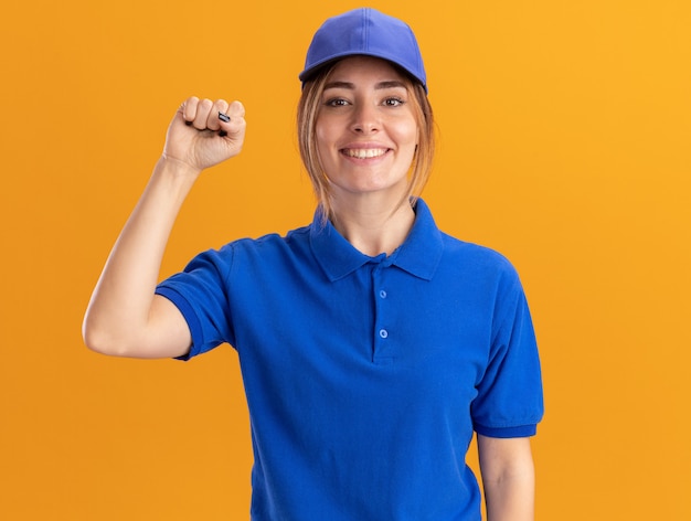 Smiling young pretty delivery woman in uniform stands with raised fist up isolated