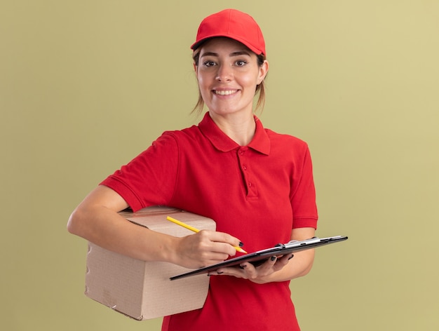 Smiling young pretty delivery woman in uniform writes on clipboard with pencil holding cardbox isolated on olive green wall