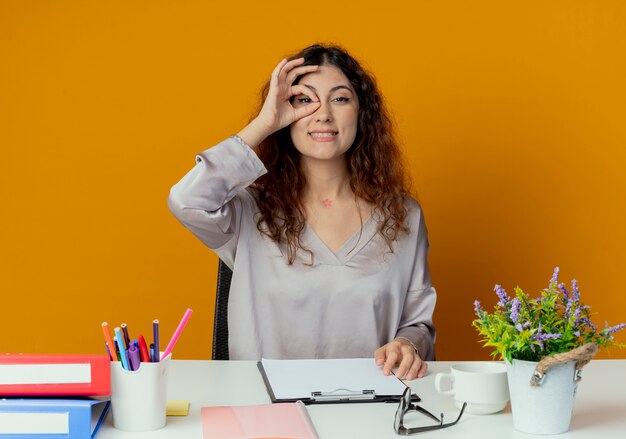 Smiling young pretty female office worker sitting at desk with office tools showing look gesture isolated on orange