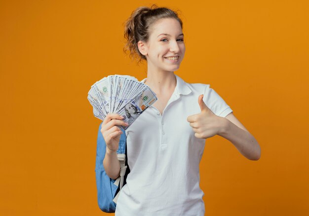 Smiling young pretty female student wearing back bag holding money and showing thumb up isolated on orange background with copy space