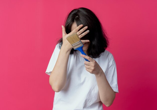 Smiling young pretty girl holding paint brush and hiding face behind hand