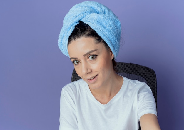 Smiling young pretty girl sitting at makeup table with makeup tools and with bath towel on head looking at camera isolated on purple background