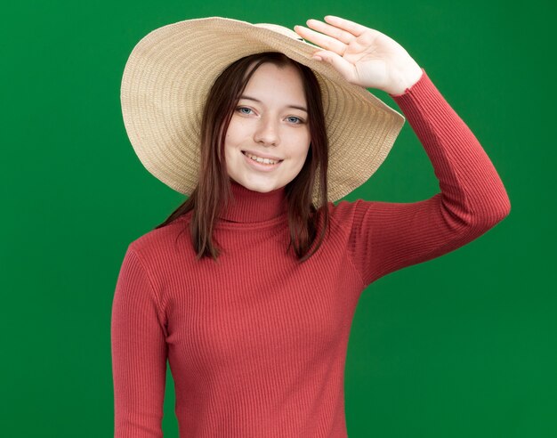 緑の壁に分離された帽子をつかむビーチ帽子を身に着けている若いかわいい女の子の笑顔