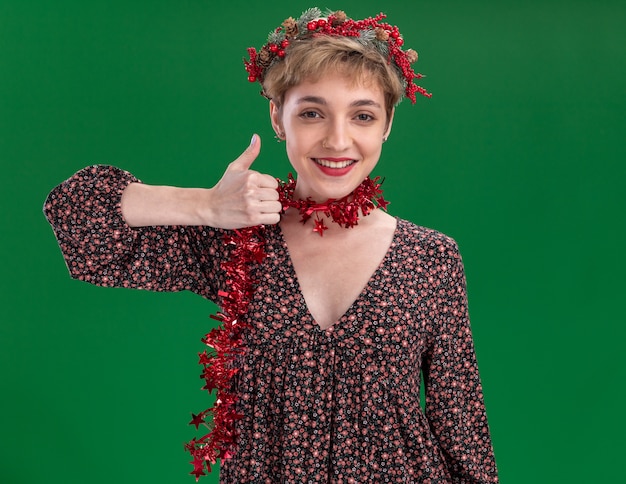 smiling young pretty girl wearing christmas head wreath and tinsel garland around neck  showing thumb up isolated on green wall