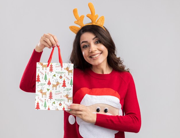 Улыбающаяся молодая красивая девушка в повязке на голову с оленьими рогами и свитере санта-клауса с рождественским подарком