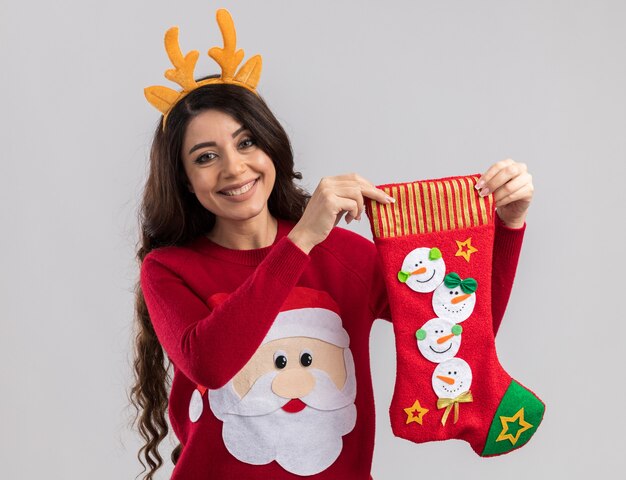 Smiling young pretty girl wearing reindeer antlers headband and santa claus sweater holding christmas stocking  isolated on white wall