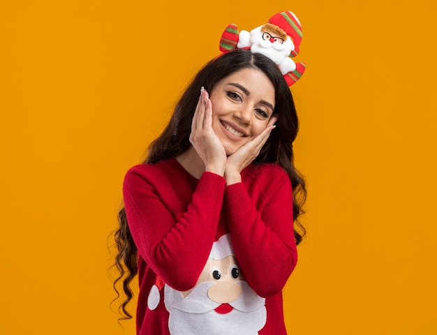 Smiling young pretty girl wearing santa claus headband and sweater  keeping hands on face isolated on orange wall with copy space