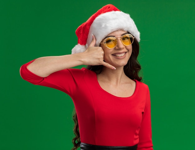 Smiling young pretty girl wearing santa hat and glasses looking at camera doing call gesture isolated on green background