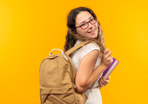 Smiling young pretty schoolgirl standing in profile view wearing glasses and back bag holding book isolated on yellow wall