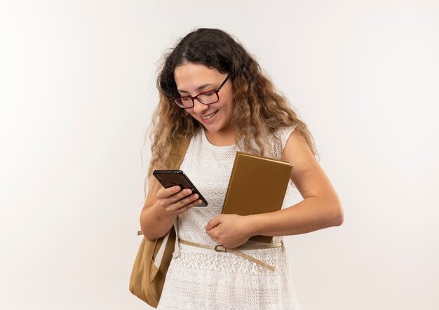 Smiling young pretty schoolgirl wearing glasses and back bag holding book using her phone isolated on wall