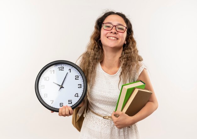 Smiling young pretty schoolgirl wearing glasses and back bag holding books and clock isolated on white wall