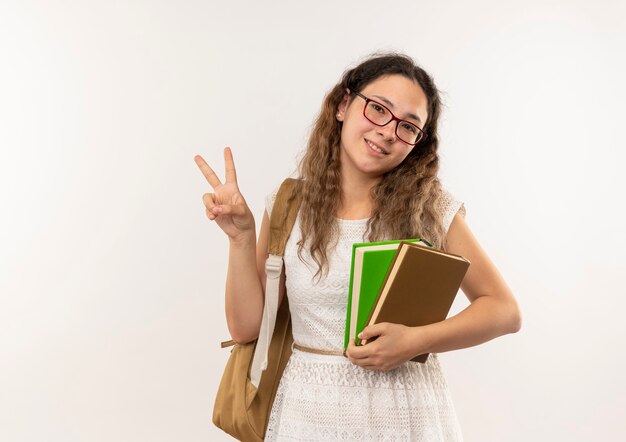 Smiling young pretty schoolgirl wearing glasses and back bag holding books doing peace sign isolated on white wall