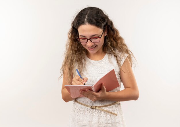 Smiling young pretty schoolgirl wearing glasses and back bag writing with pen on note pad isolated on white wall