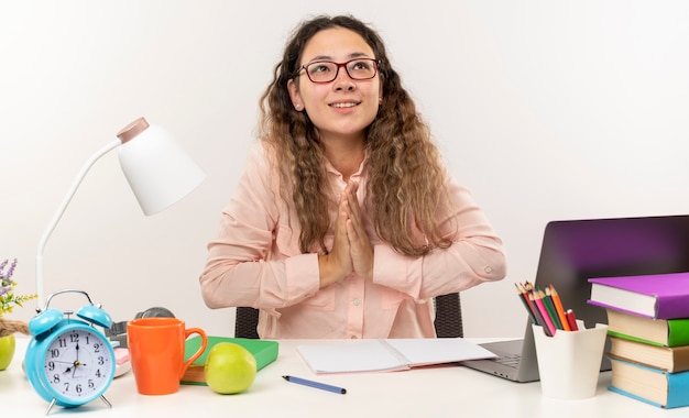 Foto gratuita sorridente giovane studentessa graziosa con gli occhiali seduto alla scrivania con gli strumenti della scuola che fa il suo lavoro mettendo le mani nel gesto di preghiera alzando lo sguardo isolato sul muro bianco