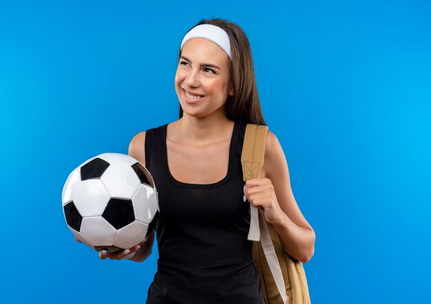 Улыбающаяся молодая симпатичная спортивная девушка с головной повязкой и браслетом и задней сумкой держит футбольный мяч и смотрит в сторону, изолированную на синем пространстве