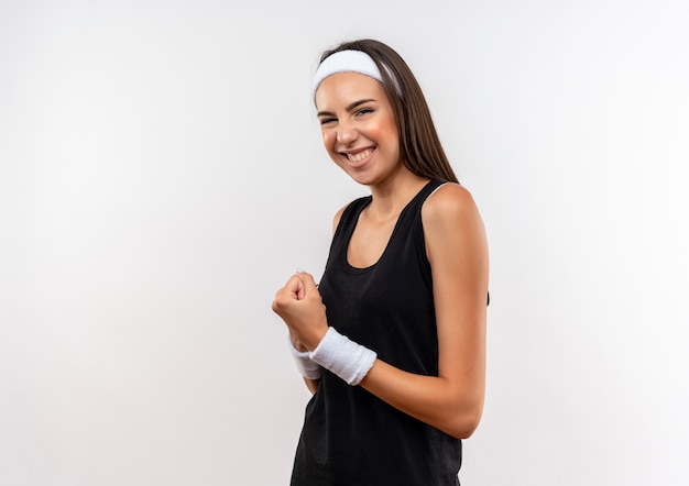 Smiling young pretty sporty girl wearing headband and wristband clenching fist on white space 