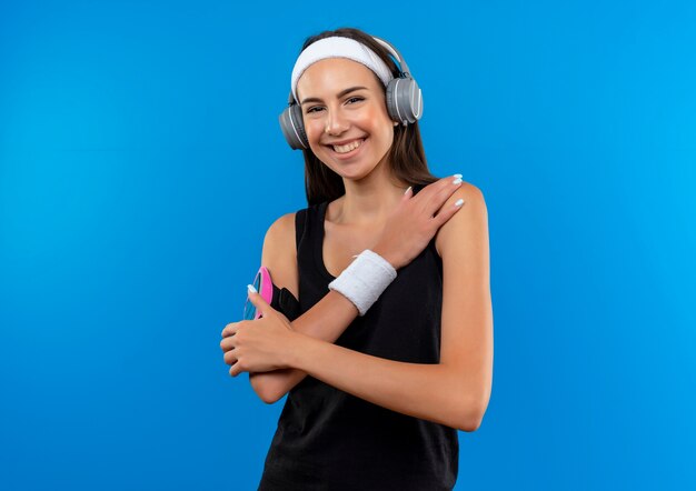 Улыбающаяся молодая симпатичная спортивная девушка с повязкой на голову и браслетом и наушниками с повязкой для телефона, положив руку на плечо, изолированную на синем пространстве