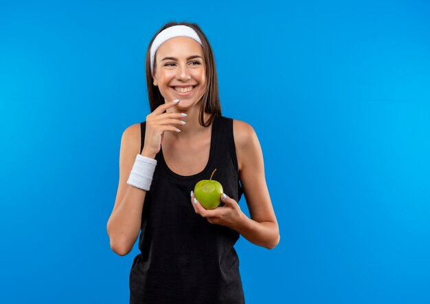 Улыбающаяся молодая симпатичная спортивная девушка с головной повязкой и браслетом, держащая яблоко, смотрит в сторону с рукой на подбородке, изолированной на синем пространстве