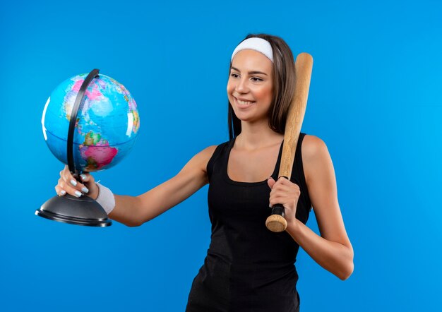 Улыбающаяся молодая симпатичная спортивная девушка с головной повязкой и браслетом, держащая бейсбольную биту и глобус, глядя на глобус, изолированный на синем пространстве
