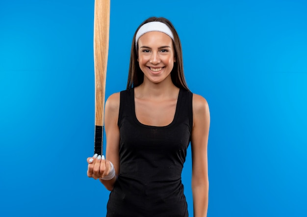 Foto gratuita sorridente giovane ragazza abbastanza sportiva che indossa la fascia e il braccialetto tenendo la mazza da baseball isolato su spazio blu