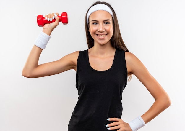 Smiling young pretty sporty girl wearing headband and wristband holding dumbbell putting hand on waist isolated on white space