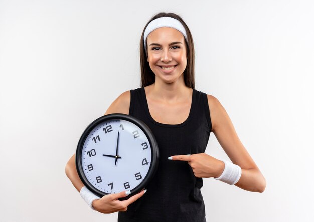 Smiling young pretty sporty girl wearing headband and wristband holding and pointing at clock on white space