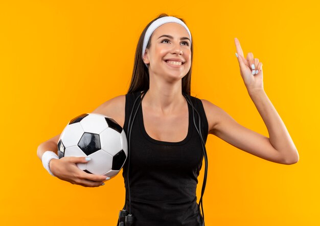 Улыбающаяся молодая симпатичная спортивная девушка с ободком и браслетом держит футбольный мяч со скакалкой на шее, изолированной на оранжевом пространстве