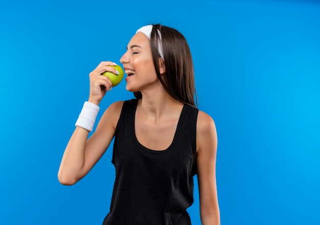 Smiling young pretty sporty girl wearing headband and wristband holding and trying to bite apple with closed eyes isolated on blue space 