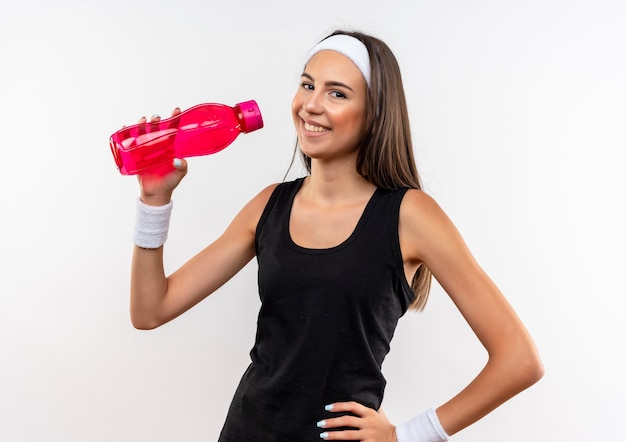 Smiling young pretty sporty girl wearing headband and wristband holding water bottle with hand on waist on white space