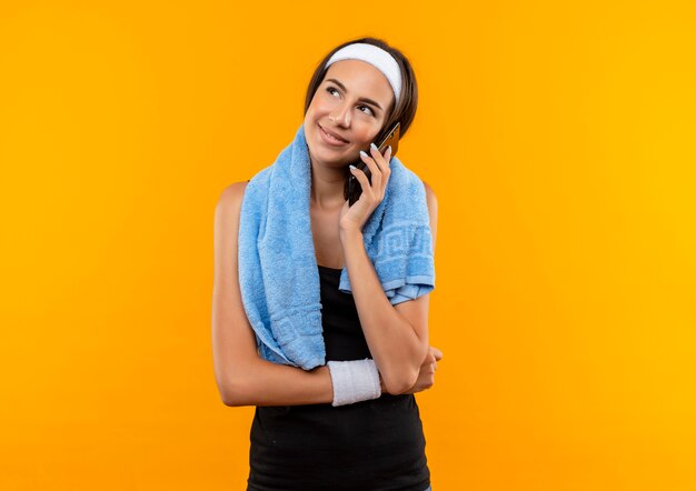 Улыбающаяся молодая симпатичная спортивная девушка с повязкой и браслетом разговаривает по телефону с полотенцем на шее на оранжевом пространстве