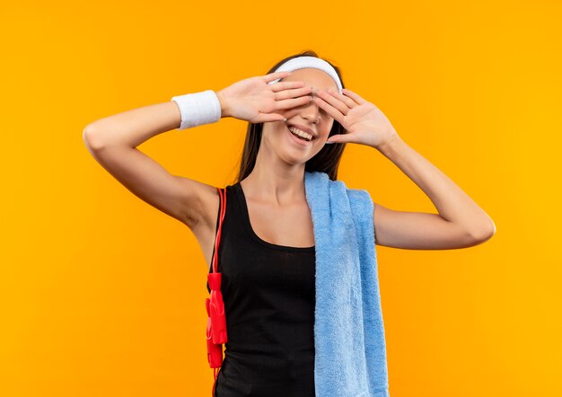 Улыбающаяся молодая симпатичная спортивная девушка с повязкой на голову и браслетом с полотенцем и скакалкой на плечах, закрывая глаза руками на оранжевом пространстве