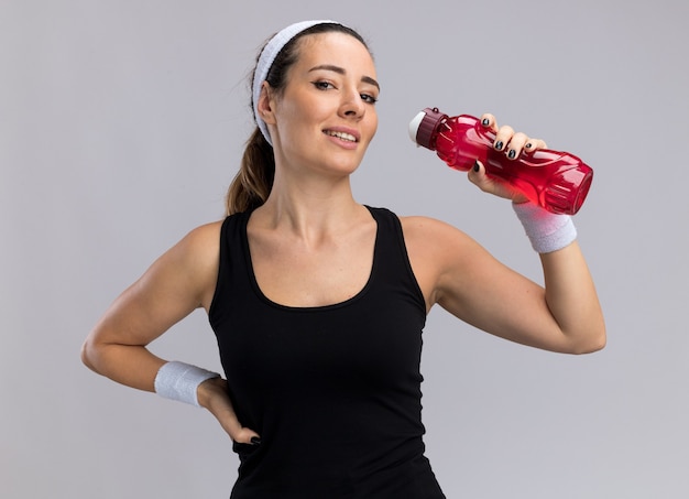 Smiling young pretty sporty girl wearing headband and wristbands holding water bottle keeping hand on waist  isolated on white wall