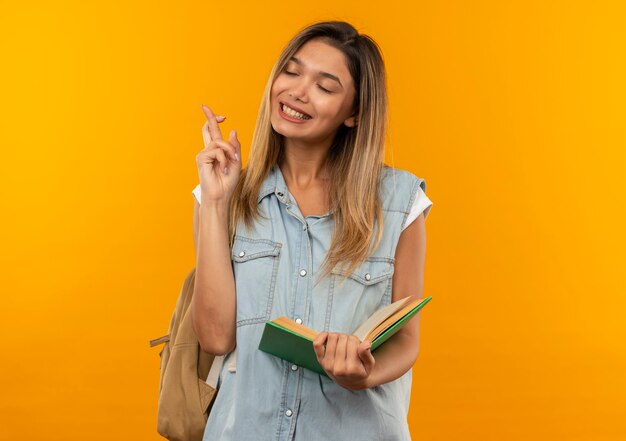 Улыбающаяся молодая симпатичная студентка в задней сумке держит открытую книгу, скрестив пальцы с закрытыми глазами, изолированными на оранжевой стене