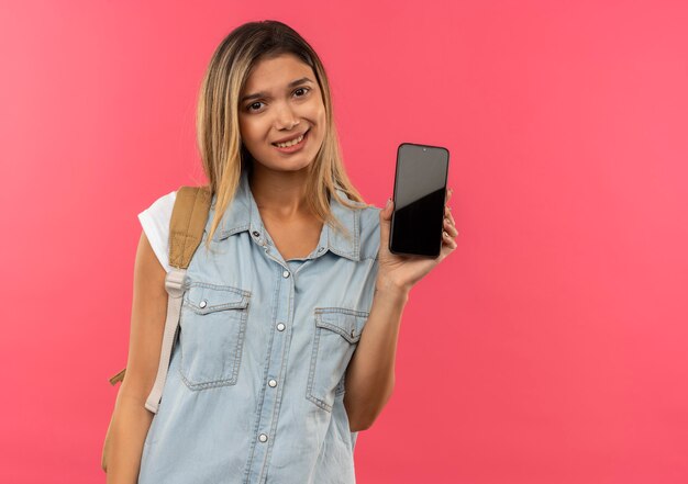 Smiling young pretty student girl wearing back bag showing mobile phone isolated on pink wall