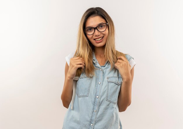 Улыбающаяся молодая симпатичная студентка в очках и задней сумке, держащая ремни задней сумки, изолирована на белой стене